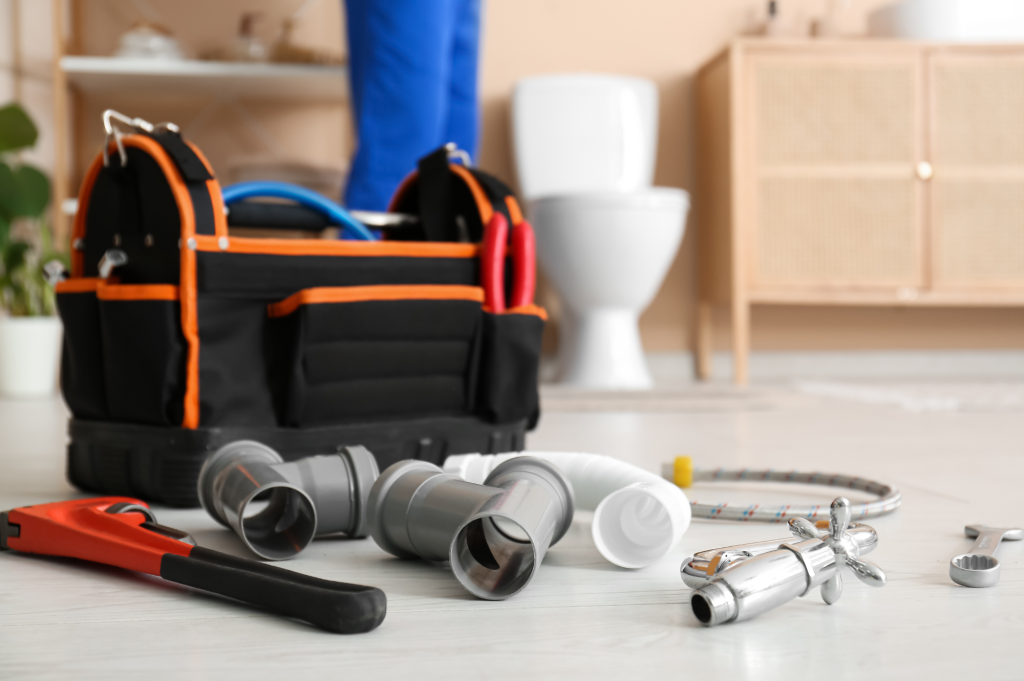 Plumbing tools and pipes on a bathroom floor with a toilet and plumber in the background.
