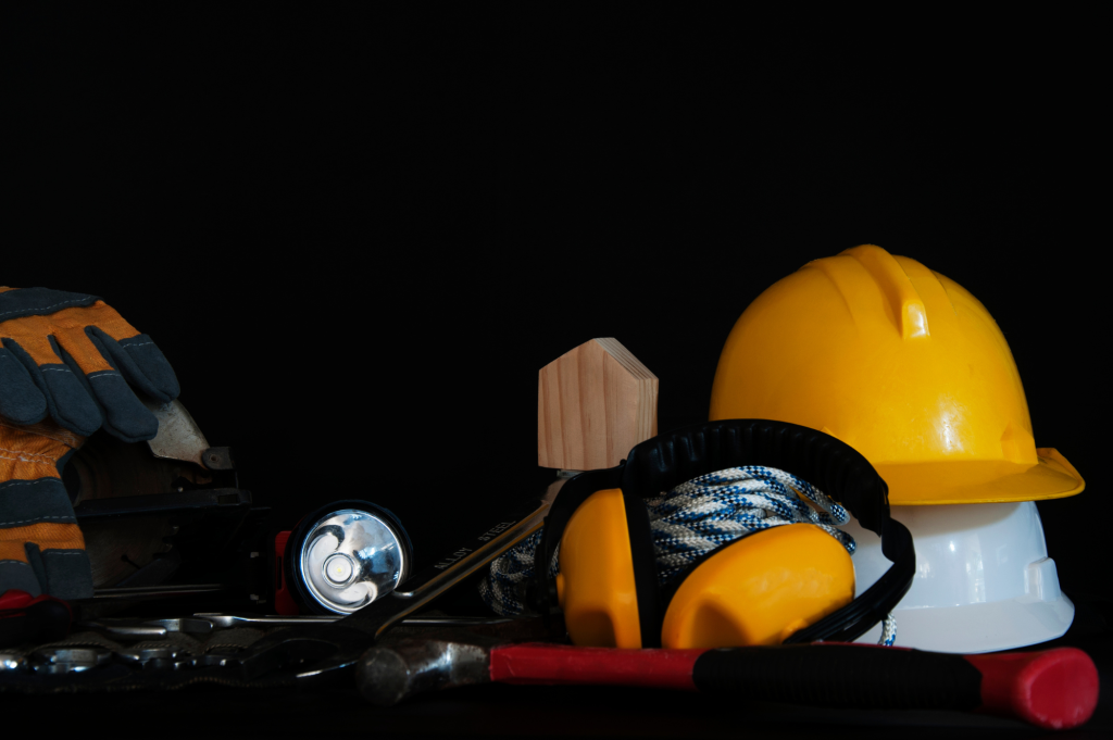Construction safety gear and tools including a yellow hard hat, protective earmuffs, and a flashlight on a dark background.