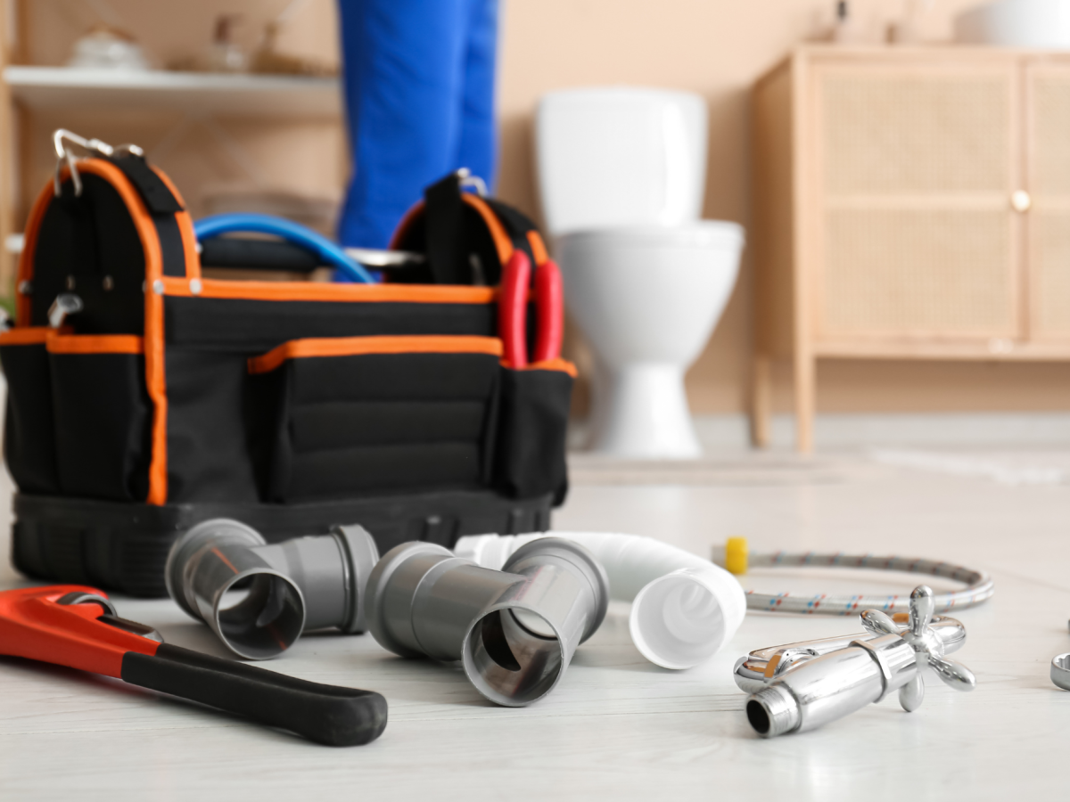 Plumbing tools and pipes on a bathroom floor with a toilet and plumber in the background.